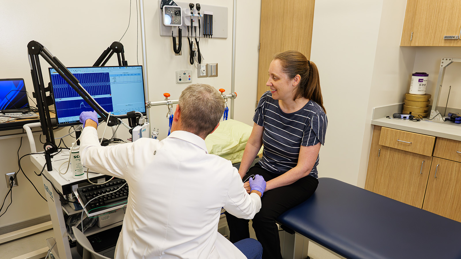 A physician evaluates Dr. Moss's muscles and nerves in an exam room.